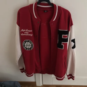Röd och vit bomberjacka från FSBN - Snygg röd och vit bomberjacka med broderade patches och text som 'World Champions' på ryggen. Jackan har klassisk college-stil med ribbade muddar, knappar framtill och kontrastärmar. Perfekt för dig som gillar sportig och trendig look.