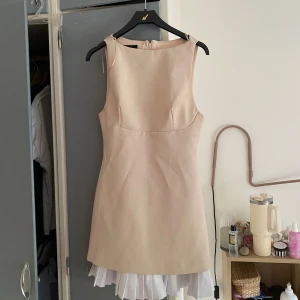 Beige klänning Zara - Supersöt beige ärmlös kortklänning med rund hals och vit plisserad kant längst ner. Klänningen har en enkel, rak passform och dragkedja i ryggen. Perfekt för dig som gillar stilrena och ljusa plagg.   