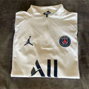 PSG half zip träningströja - Snygg vit långärmad PSG träningströja från Jordan med half zip och broderad klubbmärke på bröstet. Svarta och röda detaljer på ärmarna, tryckt logga och text på bröst och ärm. Perfekt för fotboll eller chill.🤙🤩