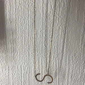 Guldfärgat halsband med S-hänge - Snyggt halsband i guldfärgad metall med en tunn kedja och ett stort S-format hänge. Perfekt för dig som vill ha en personlig touch eller gillar bokstavssmycken. Justerbar längd med karbinlås. Stilrent och enkelt att matcha med andra smycken.