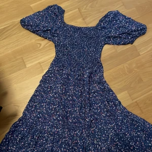 Blommig blå långklänning med puffärm - Supersöt långklänning i blått med småblommigt mönster i vitt och rött. Klänningen har smockad överdel, puffiga korta ärmar och en flowy kjol. Perfekt för sommarens alla tillfällen! Materialet känns lätt och luftigt.