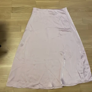 Ljusrosa satinkjol med dragkedja - Superfin ljusrosa långkjol i satin med lätt glansig finish. Kjolen har en smal passform upptill och faller snyggt ut mot nederkanten. Stängs med en diskret dragkedja i sidan. Perfekt för dig som gillar en clean och stilren look. d