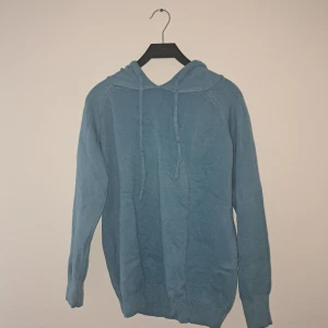 Blå stickad hoodie med huva - Mysig blå hoodie i stickat material med huva och snörning. Tröjan har långa ärmar och ribbade muddar vid ärmslut och nederkant. Perfekt för  kyliga sommardagar när du vill vara både bekväm och snygg