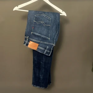 Replay Anbass slim fit jeans blå - Snygga Replay Anbass jeans i blå tvätt med slim fit passform. Klassisk femficksmodell med ljusa slitningar och kontrastsömmar. Märkespatch i läder baktill och diskret logga på bakfickan. Perfekta för dig som gillar en smal siluett och stilren look.