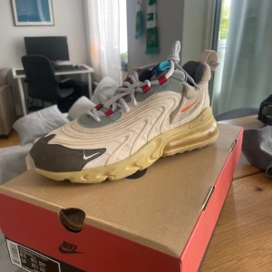 Nike Air Max 270 React Travis scott  - Nike Air Max 270 React i beige, grå och bruna toner med röda detaljer. Snygg vågig design, synlig Air Max-sula och små orange swoosh-loggor. Ovandel i syntet och textil, plös med blå detalj och snörning. Perfekta sneakers för dig som gillar streetstyle.