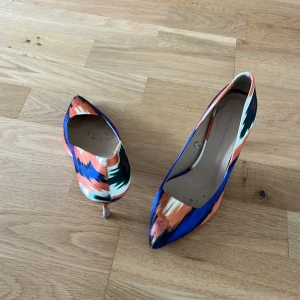 Färgglada pumps från Zara Basic - Snygga pumps från Zara Basic med spetsig tå och klack. Skorna har ett färgstarkt mönster i blått, orange, vitt och svart. Perfekta för dig som vill sticka ut med din stil. Tillverkade i textilmaterial med en elegant siluett.