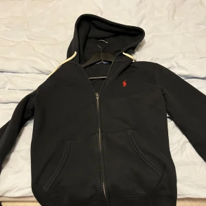 Svart hoodie från Polo Ralph Lauren XS - Svart hoodie från Polo Ralph Lauren med dragkedja och huva. Klassisk röd logga broderad på bröstet och vita snören i huvan. Mjuk bomullskänsla och kängurufickor framtill. Väldigt bra skick.