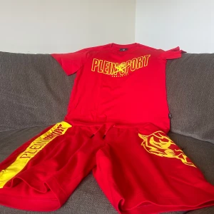 Plein Sport Set storlek XL - Snyggt set från Plein Sport med gul logga och tigertryck på ena benet. Shorts med snörning i midjan och bred gul sidopanel med röd text. Tillverkade i 100% bomull för en skön känsla. Perfekta för dig som vill sticka ut med färg och stil.