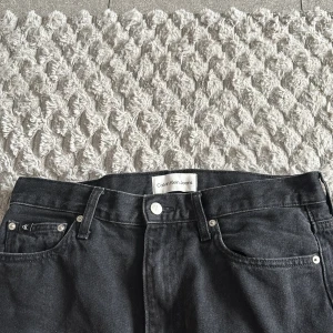Svarta high rise jeans Calvin Klein - Snygga svarta jeans från Calvin Klein med high rise och raka ben. Klassisk femficksmodell med silverfärgade detaljer och logga på bakfickan. Jeansen är i ett mjukt denimtyg och passar perfekt till en avslappnad stil.