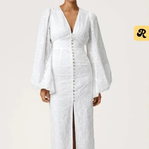 Vit långklänning med puffärmar från Adoore - Superfin vit långklänning med v-ringning, puffiga långärmar och knappar framtill. Klänningen har broderade blommor i tyget och en slits framtill som ger en elegant touch. Endas använd en gång. 