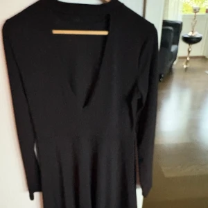 Svart långärmad klänning med v-ringning - En stilren svart klänning med lång ärm och snygg v-ringning. Modellen är enkel och tidlös med lätt utställd kjol, perfekt för dig som gillar minimalistisk stil. Materialet känns mjukt och följsamt mot kroppen.