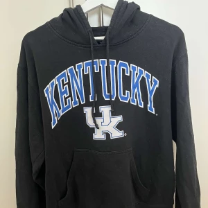 Svart Kentucky hoodie med tryck - Svart hoodie med stor KENTUCKY-text och UK-logga i blått och vitt på bröstet. Klassisk känguruficka framtill, snörning i huvan och långa ärmar. Perfekt för dig som gillar college-stil och vill ha en skön och avslappnad look.
