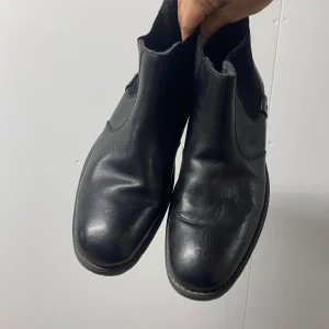 Svarta chelsea boots i skinn - Klassiska svarta chelsea boots med rund tå och resår på sidorna. Skorna har en platt sula med diskret klack och är tillverkade i slätt skinn. Perfekta för dig som gillar en stilren och tidlös look.