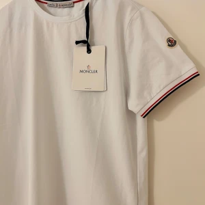 Vit Moncler t-shirt med röd/blå kant - Vit t-shirt från Moncler i slim fit-modell. Klassisk design med rund hals och korta ärmar. Ärmsluten har snygga rödblå ränder och Moncler-logga på vänster ärm. Tillverkad i mjuk bomull för en clean och fräsch look.