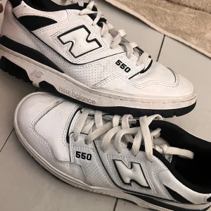 New Balance 550 vita sneakers - Säljer ett par klassiska New Balance 550 sneakers i vitt med svarta detaljer. Skorna har en chunky siluett, perforerade paneler och tydlig logga på sidan. De har snörning och platt sula, tillverkade i skinn och syntet. Perfekta för dig som gillar retrostil och streetwear.
