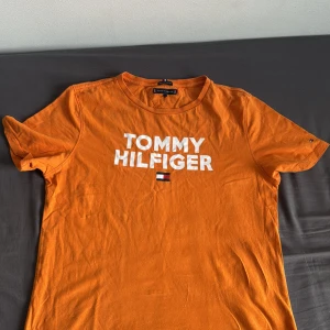 Orange t-shirt från Tommy Hilfiger - Snygg orange t-shirt från Tommy Hilfiger med vit logga och flagga på bröstet. Klassisk rund halsringning och korta ärmar. Tillverkad i 100% ekologisk bomull för en skön och mjuk känsla. Perfekt för dig som gillar färg och streetstyle.