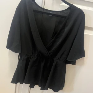 Svart omlottblus från SHEIN - Svart blus från SHEIN i storlek M med snygg omlott-skärning och v-ringning. Modellen har korta, vida ärmar och markerad midja med volangkant nedtill. Tunt och lätt material som ger en luftig känsla. Perfekt för dig som gillar stilrena och trendiga plagg.