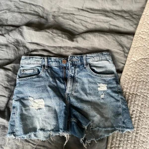 Blå jeansshorts med slitningar - Snygga blå jeansshorts med slitna detaljer och fransig nederkant. Klassisk femficksmodell med bälteshällor och dragkedja. Perfekta för varma dagar när du vill ha en avslappnad och trendig look. Storlek 146-140