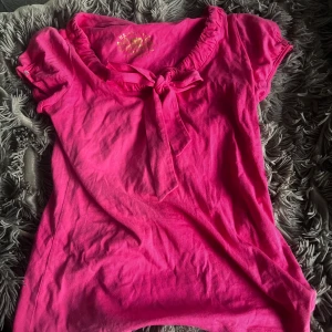 Rosa topp med knyt från U.S. Polo Assn. - Snygg rosa topp från U.S. Polo Assn. med korta ärmar och en söt knytning vid halsen. Toppen har en rundad krage med rynkade detaljer och är gjord i mjuk bomull, perfekt för en chill och färgglad look.