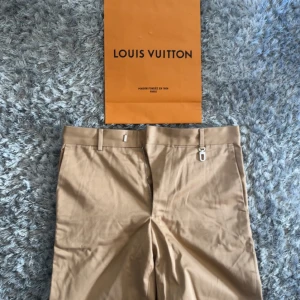 Beige kostymbyxor från Louis Vuitton - Snygga beige kostymbyxor från Louis Vuitton med exklusiva metalldetaljer och diskret logga på bältet. Byxorna har klassisk passform, hällor för bälte och är tillverkade i bomullsmix. Perfekta för en clean och stilren look.