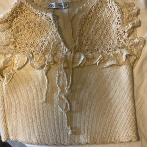 Beige stickad topp med spets från Zara - Superfin beige stickad topp från Zara med spetsdetaljer och volanger upptill. Toppen har knytband framtill och är ärmlös med en croppad passform. Perfekt för dig som gillar romantiska och unika detaljer i din stil.