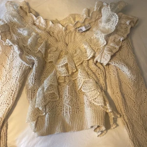 Beige stickad blus med spetsvolang zara - Supersöt beige stickad blus från H&M med breda spetsvolanger längs ringningen och axlarna. Långärmad och med v-ringad hals. Perfekt för dig som gillar romantisk och vintage-inspirerad stil.