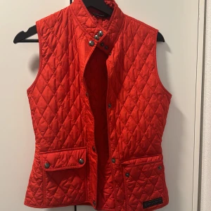 Röd quiltad dunväst från Belstaff - Snygg röd quiltad dunväst från Belstaff med hög krage och tryckknappar. Västen har två stora fickor framtill med lock och knappar samt ett diskret Belstaff-märke nedtill. Pris kan diskuteras vid snabb affär 