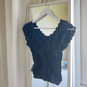Svart volang topp med v-ringning - Supersnygg svart topp med djup v-ringning och stora volanger längs halsen och axlarna. Stickad känsla och ärmlös design som ger en cool och trendig look. Perfekt att styla med jeans eller kjol för en extra touch.