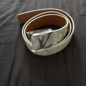 Louis V bälte – klassisk monogramstil - Lyft din outfit med detta exklusiva bälte i brun läderlook och klassiska monogramdetaljer. Spännet har en stilren design i metall som ger ett lyxigt intryck – perfekt för både vardags- och festlooken.