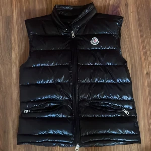 Svart dunväst från Moncler - Snygg svart dunväst från Moncler med glansig finish och klassisk logga på bröstet. Västen har hög krage, två fickor med dragkedja och quiltad design. Perfekt att slänga över en hoodie för en clean och trendig look. Bara fråga om funderingar! Pris kan diskuteras.