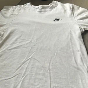 Vit Nike t-shirt med logga - Klassisk vit t-shirt från Nike med svart broderad logga på bröstet. Enkel och stilren design med rund hals och korta ärmar. Tillverkad i mjuk bomull som känns skön mot huden. Perfekt för en clean och sportig look.