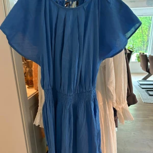 Blå kortklänning med knyt i ryggen - Supersöt blå klänning med kort längd och vida ärmar. Klänningen har smock i midjan för en snygg passform och knytning i ryggen som ger en öppen rygg-detalj. Perfekt för varma dagar och enkel att styla med sneakers eller sandaler.