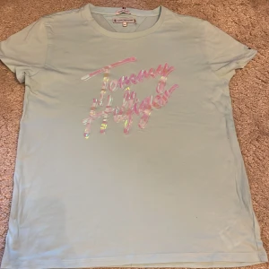 Ljusblå Tommy Hilfiger t-shirt - Snygg ljusblå t-shirt från Tommy Hilfiger med holografiskt tryck på bröstet. Tillverkad i mjuk ekologisk bomull, klassisk rund hals och korta ärmar. Perfekt för dig som gillar stilrena plagg med en cool twist.