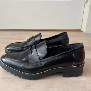 Svarta chunky loafers i skinn - Snygga svarta loafers med chunky sula och klassisk penny-detalj. Skorna är tillverkade i blankt skinn och har en markerad sula med grovt mönster för extra edge. Perfekta för dig som vill ha en stilren men trendig look.