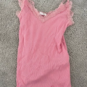 Rosa ribbad linnetopp med spets - Supersöt rosa linnetopp med ribbad struktur och fina spetsdetaljer längs v-ringningen och axelbanden. Perfekt för att ge outfiten en romantisk touch. Tunn och lätt, passar perfekt till varma dagar.