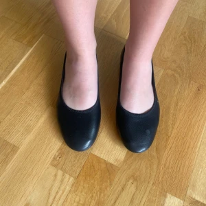 Svarta ballerinaskor  - Klassiska svarta ballerinaskor från 157 i storlek 38. Skorna har rund tå, platt sula och är gjorda i syntetmaterial med lätt struktur. Perfekta för en clean och enkel look. Sulan har ett hjärtmönster för extra detalj. Rosetten avklippt 