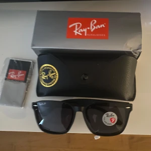 Svarta Ray-Ban Wayfarer solglasögon - Klassiska svarta Ray-Ban Wayfarer solglasögon med polariserade mörka glas. Snygg och tidlös design med robust plastbåge. Kommer med originalfodral och putsduk. Perfekta för dig som vill ha en ikonisk look och skydda ögonen mot solen.