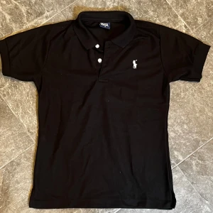 Svart pikétröja från Ralph Lauren - Klassisk svart pikétröja från Ralph Lauren med vit broderad logga på bröstet. Tröjan har korta ärmar, krage och knäppning med två vita knappar. Perfekt för en stilren och sportig look.