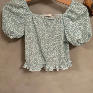 Blå leopardmönstrad blus KappAhl - Supersöt blus från KappAhl i ljusblått med vitt leopardmönster. Modellen är croppad med puffiga korta ärmar, smockad rygg och volangkant nedtill. Perfekt för dig som gillar trendiga detaljer och djurmönster. Skriv vid frågor!💗