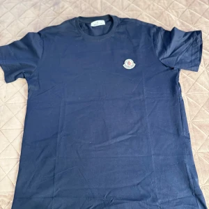 Mörkblå Moncler t-shirt i bomull - Snygg mörkblå t-shirt från Moncler med klassisk rund hals och korta ärmar. T-shirten har en broderad Moncler-logga på bröstet och är tillverkad i 100% bomull för en skön och mjuk känsla. Perfekt för dig som gillar stilrena och exklusiva plagg. Storlek s i man storlek
