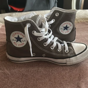 Bruna Converse Chuck Taylor All Star - Klassiska höga Converse Chuck Taylor All Star i brun canvas med vita gummidetaljer och snörning. Skorna har rund tå, vit sula med svart rand och ikonisk logga på sidan. Perfekta för en avslappnad och cool stil.