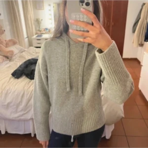 Grå stickad hoodie med snörning - Mysig grå stickad hoodie med huva och snörning. Tröjan har långa ärmar och breda ribbade muddar vid ärmslut och nederkant. Skriv för funderingar eller fler bilder