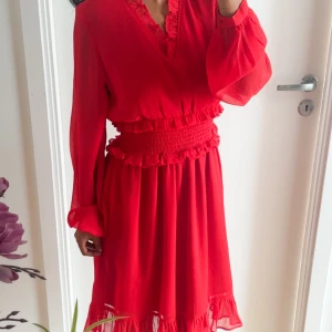 Röd volangklänning från NA-KD - Superfin röd klänning från NA-KD med v-ringning, långa ärmar och smock i midjan. Klänningen har volangdetaljer vid halsringning, midja och nederkant. Lätt och luftig med transparenta partier vid ärmar och fåll. Perfekt för sommaren! Priset kan diskuteras 