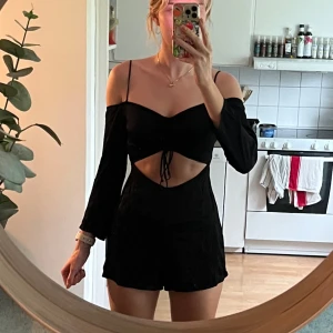Svart byxdress med cut out  - En jättefin kort byxdress med långa ärmar och tunna axelband. Snygg cut out-detalj och snörning framtill. Den har en dragkedja i sidan och är i sidenliknande material. Tycker verkligen om denna men säljer då den inte kommer till användning, fint skick!