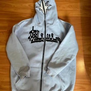 Ljusblå hoodie med tryck och huva - Säljer en ljusblå hoodie i fleece med svart och vitt tryck framtill och unik stjärndetalj på huvan. Tröjan har dragkedja hela vägen, fickor och ribbade muddar. Perfekt för dig som gillar streetwear och vill sticka ut.