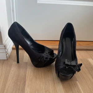 Svarta pumps med rosett och platå - Snygga svarta pumps med hög klack och platåsula. Skorna har öppen tå och en dekorativ rosett framtill. Materialet är matt och känns som mocka, med detaljer i blankt syntet.