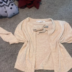 Beige kofta med rosett från 157, XS - Supermjuk beige kofta från 157 i storlek XS. Koftan har en snygg rosettdetalj vid halsen och är öppen framtill. Perfekt för lager-på-lager och chill dagar. Långärmad och i ett tunt, skönt material som känns riktigt nice mot huden.