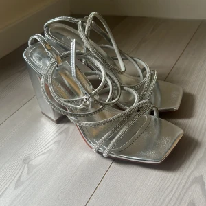 Silvriga sandaler med blockklack - Glänsande silvriga sandaler med fyrkantig tå och höga blockklackar. Skorna har tunna, glittriga remmar som korsar över foten och knyts runt vristen. Perfekta för att sticka ut och ge en futuristisk vibe till din outfit.
