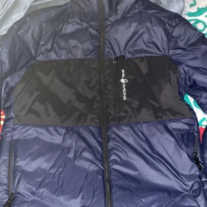 Blå och svart Sail Racing pufferjacka - Säljer en blå och svart pufferjacka från Sail Racing med huva och hel dragkedja. Jackan har en sportig look, bröstficka med dragkedja och logga på bröstet. Materialet är syntet och jackan är perfekt för kyliga dagar. Snygg och funktionell design för dig som gillar streetwear.