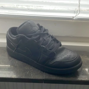 Svarta nike Air Jordan 1 Low sneakers - Svarta Nike Air Jordan 1 Low sneakers i skinn med klassisk siluett och perforerad tå. Snyggt helsvart utförande med snörning och diskret Jumpman-logga på plösen. Perfekta för dig som gillar streetstyle och vill ha en ikonisk sneaker i garderoben. Men det kommer in lite vatten under skorna.